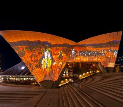 The Sydney Opera House sails at night, illuminated with an Australian outback landscape in orange and browns with a blue sky.